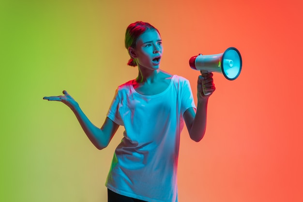 Schreeuwen met megafoon, luidspreker. het portret van het jonge kaukasische meisje op de achtergrond van de gradiëntgroen-oranje studio in neonlicht. concept van jeugd, menselijke emoties, gezichtsuitdrukking, verkoop, advertentie.