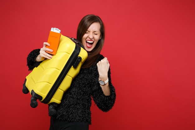 Schreeuwende jonge vrouw in zwarte vacht trui met gesloten ogen doen winnaar gebaar houden koffer, paspoort instapkaart ticket geïsoleerd op rode achtergrond. Mensen oprechte emoties, lifestyle concept.