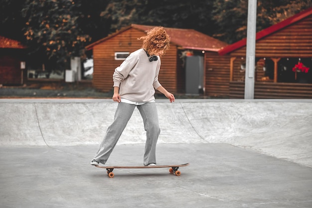 schuiven. Meisje met golvend haar met koptelefoon op haar nek op skateboard rijden op speciale ondergrond in park in de middag