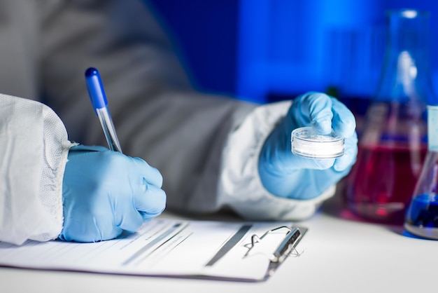 Photo science, chemistry, medicine and people concept - close up of young scientist with chemical sample taking notes on clipboard and making test or research in laboratory