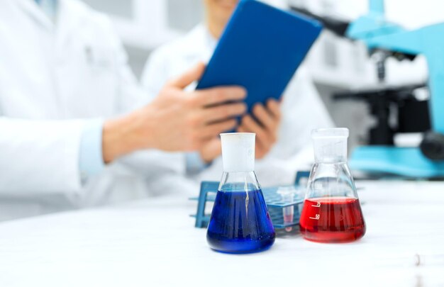 science, chemistry, technology, biology and people concept - close up of scientists with glass tes flasks and tablet pc computer on table in lab