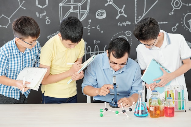 Science teacher looking through microscope, students standing around him and writing in notebook