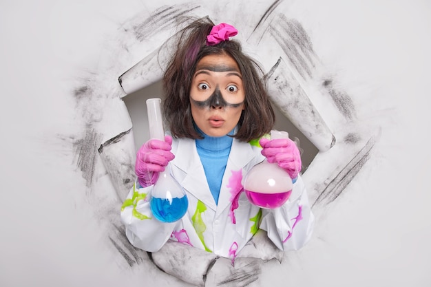 Science worker has surprised expression stares bugged eyes at camera holds two flasks with colorful liquid makes chemical experiment in scientific laboratory poses through hole in paper 