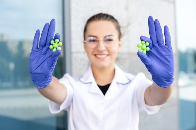 Фото Концепция науки, химии, биологии и медицины - улыбающаяся европейская девушка-медик в белом халате, очках и синих резиновых перчатках, крупным планом демонстрирует молекулы на протянутых руках на открытом воздухе.