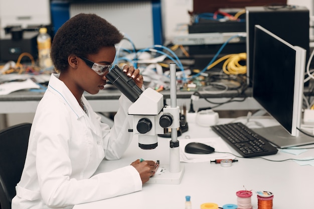 Scienziata donna afroamericana che lavora in laboratorio con strumenti tecnologici elettronici e ricerca al microscopio e sviluppo di dispositivi elettronici per colore donna nera