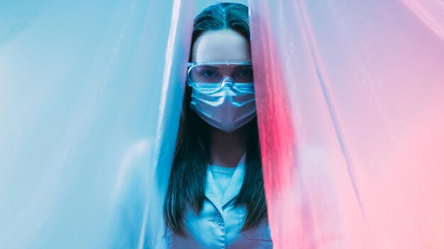 Scientist portrait Medical laboratory Clinical research Female technician doctor in protective face mask goggles in pink blue neon light isolated behind plastic curtain