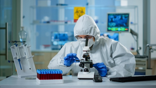 Scientist in ppe suit making adjustments and looking through laboratory microscope, typing on pc. Chemist in coverall working with various bacteria, tissue blood samples for antibiotics research