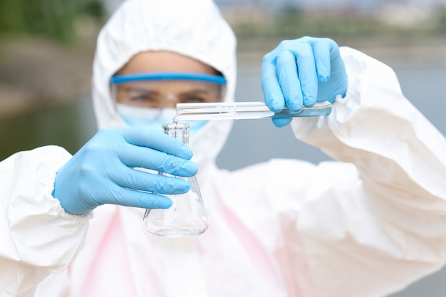 Scientist in protective suit and goggles in takes of water samples in test tube from lake