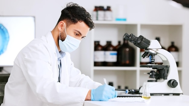 Scientist and research or medical engineer doing experiments to create a cure in a lab while wearing a mask Healthcare professional working with science equipment and writing notes in a laboratory