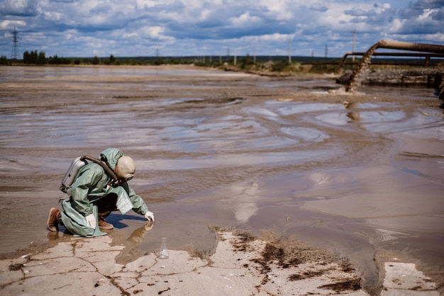 A scientist takes a sample of water from a river after the release of chemical waste A man in a respirator and a green protective suit from radiation Water analysis experts Ecological catastrophy