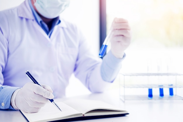 Scientist in white coat holding and examining test tube with reagent making notes of his research in laboratory