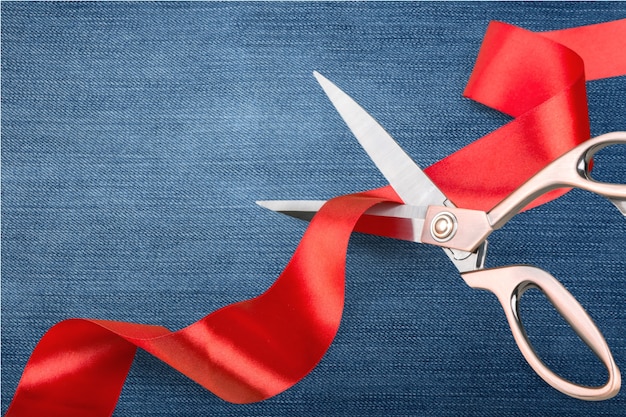 Scissors cutting red ribbon, close-up view on blue background