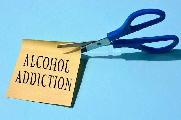 Photo scissors that cut yellow notepad with alcohol addiction text on a blue background. alcoholism concept