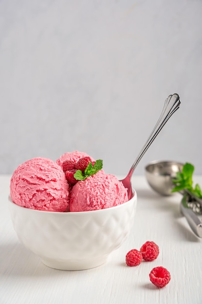 Scoops of raspberry ice cream with red berries and mint leaf served in bowl with spoon on table