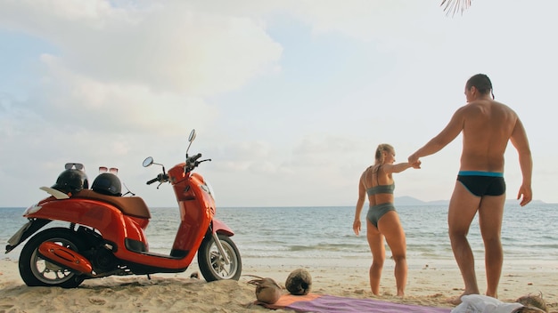 Scooter road trip. lovely couple on red motorbike in white\
clothes on sand beach. just married people kiss hugs walking near\
the tropical palm trees, sea. wedding honeymoon by ocean.\
motorcycle rent.