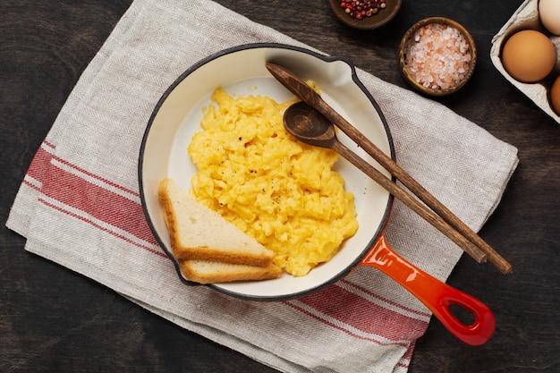 Scrambled eggs for breakfast on red old frying pan, dark wooden table wall. Traditional English breakfast. Top view with cope space.