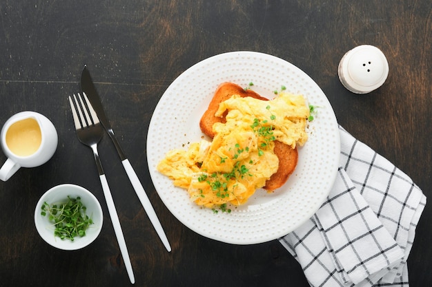 Photo scrambled eggs fluffy and buttery scrambled eggs on bread with microgreen radish and hollandaise sauce on white plate over dark old wooden background homemade breakfast or brunch meal top view