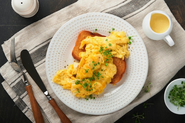 Uova strapazzate uova strapazzate soffici e burrose su pane con ravanello microgreen e salsa olandese su piatto bianco su sfondo di legno vecchio scuro colazione o brunch fatti in casa vista dall'alto