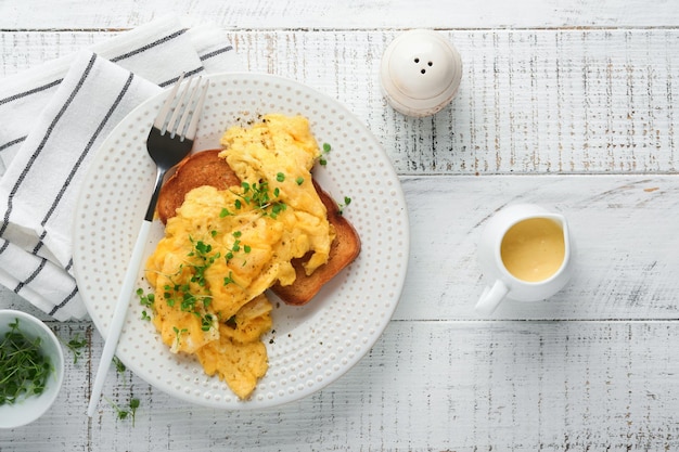 Scrambled Eggs Fluffy and Buttery scrambled eggs on bread with microgreen radish and hollandaise sauce on white plate over white wooden background Homemade breakfast or brunch meal Top view