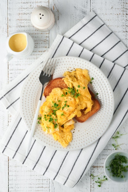 Scrambled eggs fluffy and buttery scrambled eggs on bread with\
microgreen radish and hollandaise sauce on white plate over white\
wooden background homemade breakfast or brunch meal top view