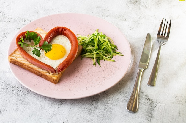 Scrambled eggs in the form of heart on a white plate with sausages, sourdough toast isolated on white marble background. Homemade food. Tasty breakfast. Selective focus. Hotizontal photo.