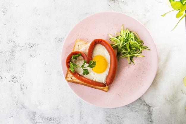 Scrambled eggs in the form of heart on a white plate with sausages, sourdough toast isolated on white marble background. Homemade food. Tasty breakfast. Selective focus. Hotizontal photo.