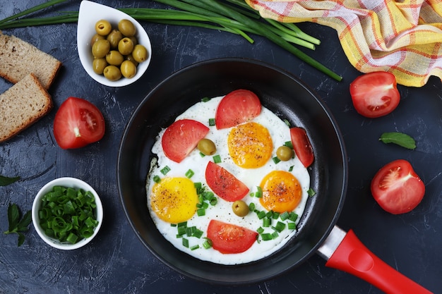 Scrambled eggs with tomatoes and green onions in a frying pan on a dark surface, on the table are olives, bread and tomatoes