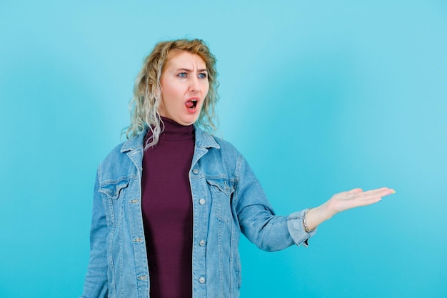 Screaming blonde woman is looking away by raising up her hand on blue background