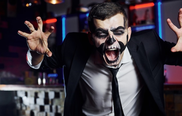 Screaming to the camera. Portrait of man that is on the thematic halloween party in scary skeleton makeup and costume.