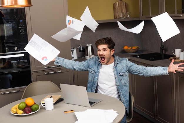 Screaming freelancer throwing documents while working with laptop on kitchen table