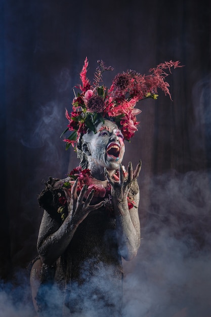 A screaming girl smeared with clay in a cemented dress. The model has a headdress made of flowers. Smoke from behind.