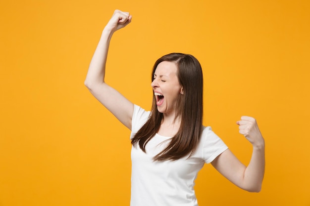 Screaming joyful young woman in white casual clothes keeping eyes closed, clenching fists like winner isolated on yellow orange wall background in studio. People lifestyle concept. Mock up copy space.