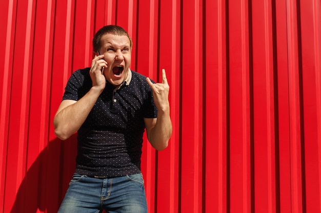 Uomo di grido con emozioni pazze usando il suo cellulare sulla parete rossa