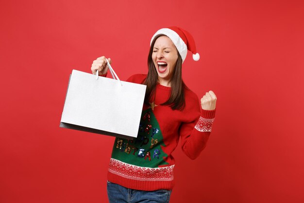 Screaming Santa girl with closed eyes clenching fist like winner, holding packages bags with purchases after shopping isolated on red background. Happy New Year 2019 celebration holiday party concept.