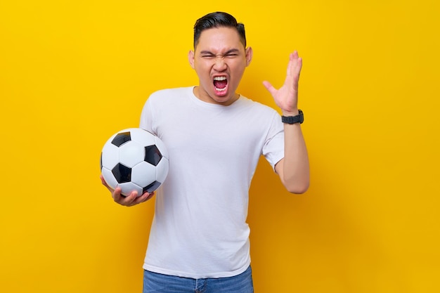 Screaming upset angry young asian man football fan in a white\
tshirt holding a soccer ball and yelling raised hand isolated on\
yellow background people sport leisure lifestyle concept