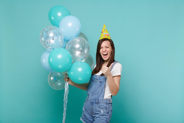 Giovane donna urlante in abiti in denim, cappello di compleanno che mostra il pollice in su per celebrare, tenendo palloncini d'aria colorati isolati su sfondo blu turchese. festa di compleanno, concetto di emozioni della gente.