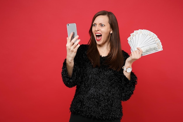 Screaming young woman in fur sweater looking on mobile phone hold fan of money in dollar banknotes cash money isolated on red background. People sincere emotions lifestyle concept. Mock up copy space.