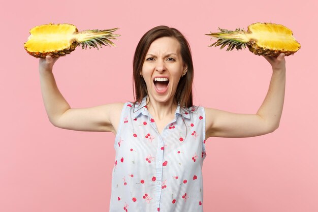 Screaming young woman in summer clothes spreading hands with halfs of fresh ripe pineapple fruit isolated on pink pastel background. People vivid lifestyle, relax vacation concept. Mock up copy space.