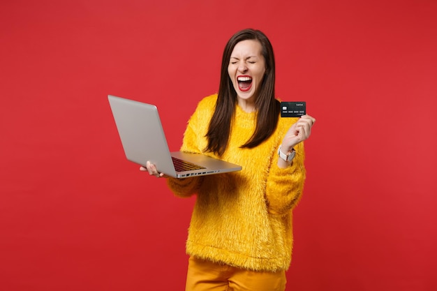 Screaming young woman in yellow fur sweater keeping eyes closed, hold credit bank card, laptop pc computer isolated on red background. People sincere emotions, lifestyle concept. Mock up copy space.