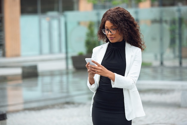 Scrolling phone chatting Africanamerican businesswoman in office attire smiling