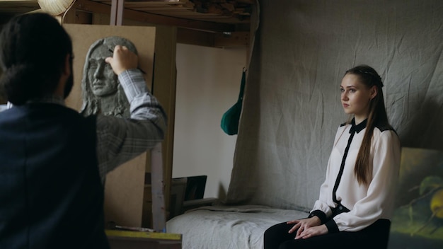 Скульптор создает скульптуру человеческого лица на холсте, пока молодая женщина позирует ему в художественной студии