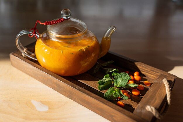 Sea buckthorn tea with mint in a transparent teapot on a wooden background in the rays of sunlight