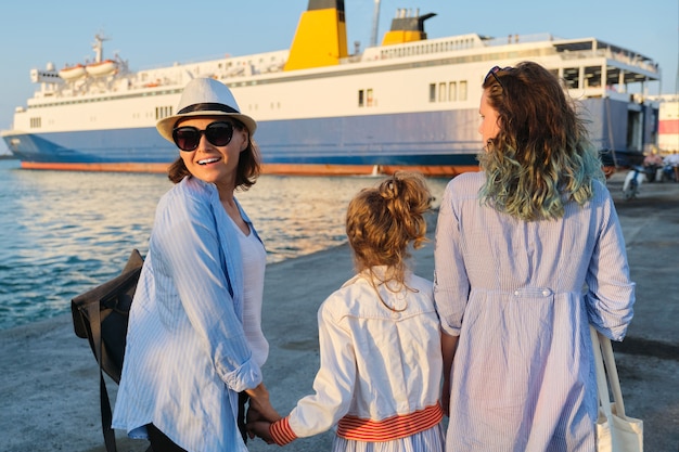 Sea family vacation, mother and daughters in the seaport holding hands looking at the ferry