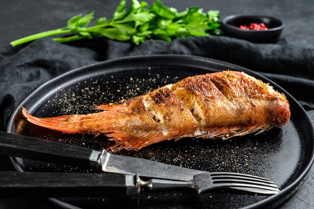 Sea perch baked served on a round plate with parsley. Black background. Top view.