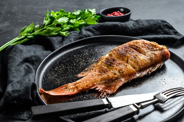 Sea perch baked served on a round plate with parsley. Black background. Top view.