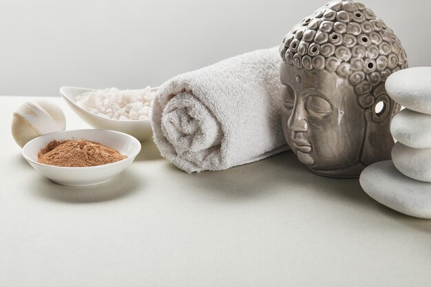 Морская соль и глиняный порошок в мисках, хлопковые полотенца, камни, бомба для ванны и фигурка Будды на белом
