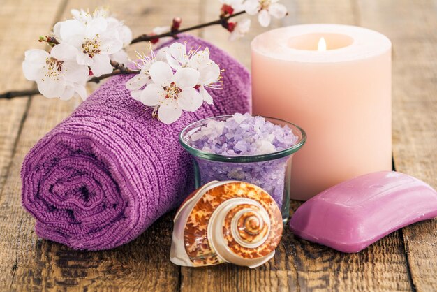 Sea salt in glass bowl with towel, sea shell, soap for bathroom procedures and burning candle with flowering branch of apricot tree on wooden background. Spa products and accessories.