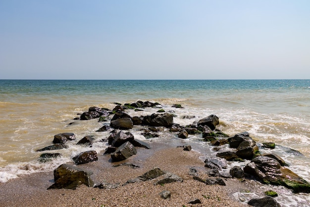 Морское побережье с камнями, волны чистой голубой воды и голубое небо на заднем плане. Побережье красивого летнего моря. Сезон путешествий и отпусков.