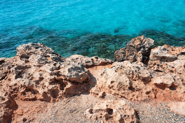Sea shore with rocks and clear transparent sea water. Natural marine background. Blue ocean wallpaper, sea wave on sunshine day. Crystal clear water and orange cliffs of the tropical sea