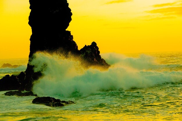 Sea waves splashing on rocks against sky during sunset